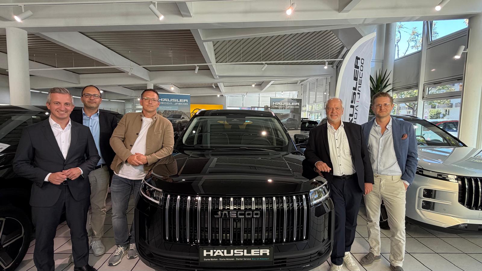 Fünf Vertreter von OMODA & JAECOO und dem Autohaus Häusler vor einem schwarzen JAECOO im Showroom in München